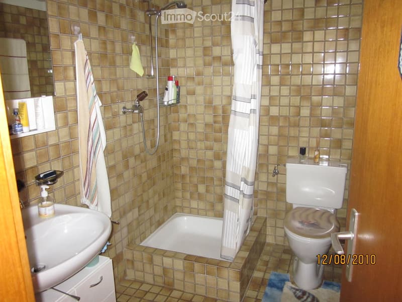 A small bathroom with beige tiled walls and floor, featuring a sink, toilet, bathtub, and shower area with a curtain. A towel hangs on the wall, and various toiletries are placed on the sink and bathtub ledge.