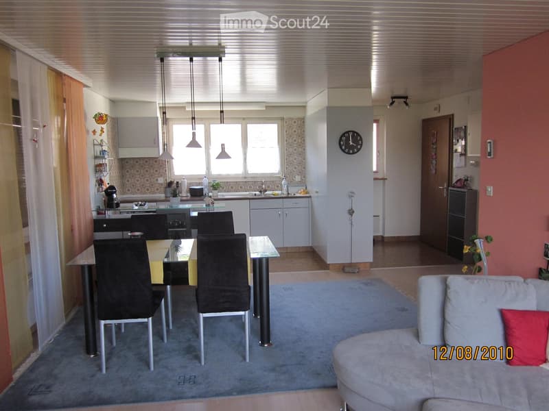 A modern, open-plan living and dining area with a grey sofa, a glass dining table with black chairs, a kitchen area with cabinets and appliances, and a clock on the wall.