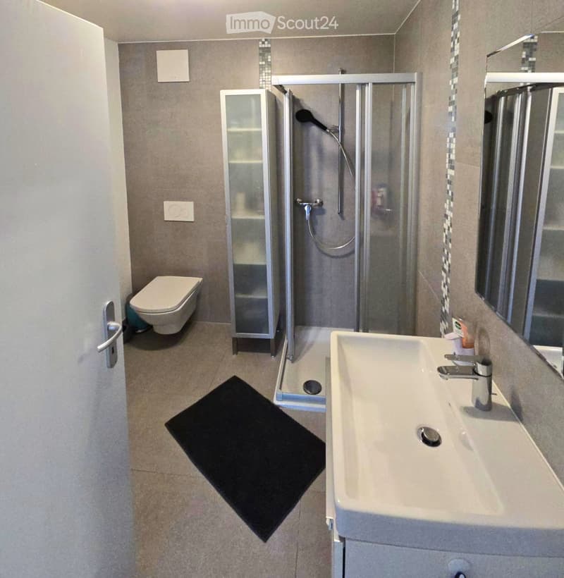 Modern bathroom with a toilet, sink, mirror, glass shower enclosure, and a small cabinet. Features a black bath mat on the floor.