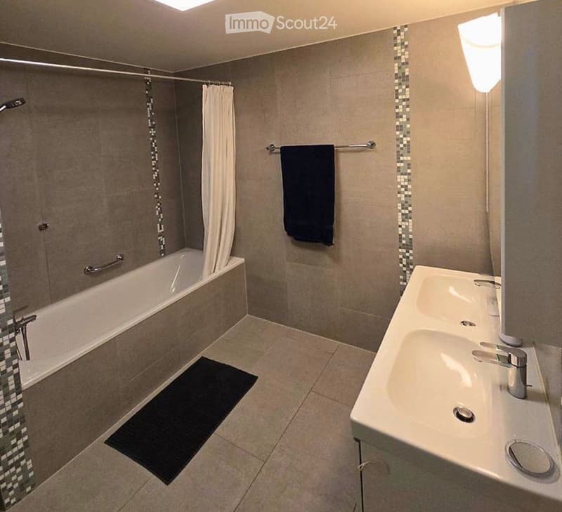 Modern bathroom with a bathtub, double sink, towel rack, and a dark towel. The walls are tiled with a decorative border, and the floor has a dark bath mat.