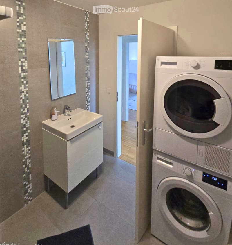 Modern bathroom with a sink, mirror, and stacked washing machine/dryer unit. The sink has a modern faucet and a small cabinet underneath.