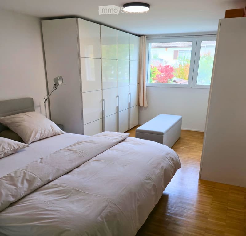 A modern bedroom with a large double bed, pillows, a wardrobe, a small stool, and a floor lamp. The room has wooden flooring and large windows with curtains.