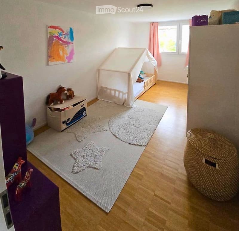 Cozy children's bedroom with a wooden floor, a toy bed with a canopy, a wicker chair, a toy box, and a colorful painting on the wall.