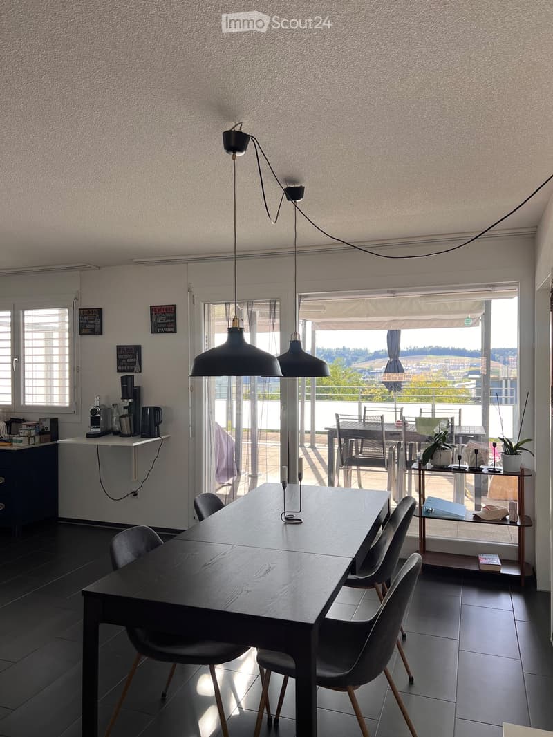 Modern dining area with a dark wooden table, six chairs, pendant lamps, a small kitchen counter with a coffee machine, and a view to a balcony with outdoor furniture.
