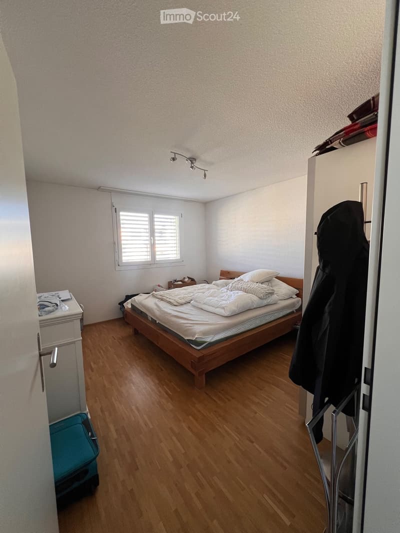 A small bedroom with wooden flooring, a bed with white bedding, a nightstand, and a coat hanging on the door.