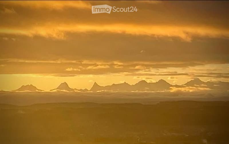 A panoramic view of a mountain range under a dramatic, golden sunset sky. The mountains are silhouetted against the warm, orange and yellow hues of the clouds.