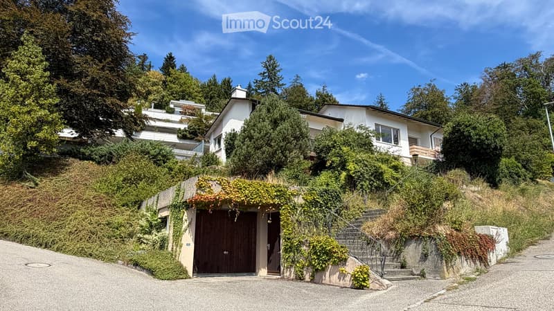 A two-story house with white walls, multiple windows, and a garage entrance, surrounded by lush greenery and trees.