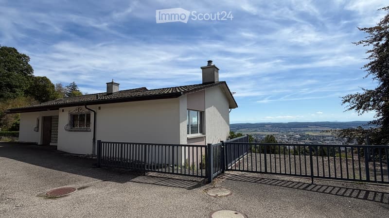 Single-story house with white exterior, dark roof, and metal gate, surrounded by greenery and offering a scenic view of the valley.
