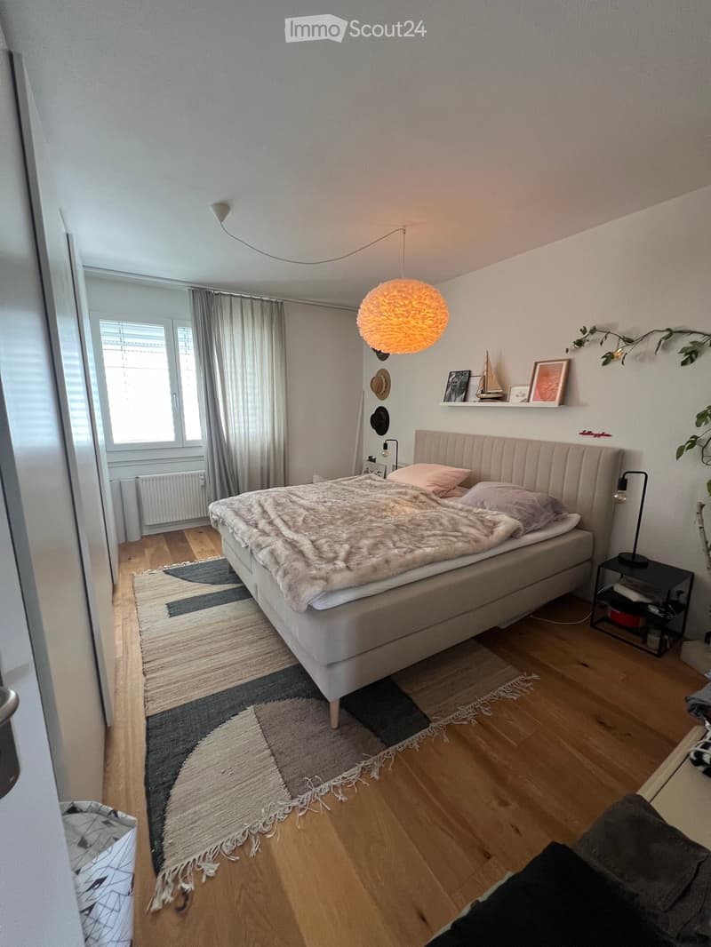 Cozy bedroom with a large bed, nightstand, and decorative shelf. Features wooden flooring, a patterned rug, and modern lighting.