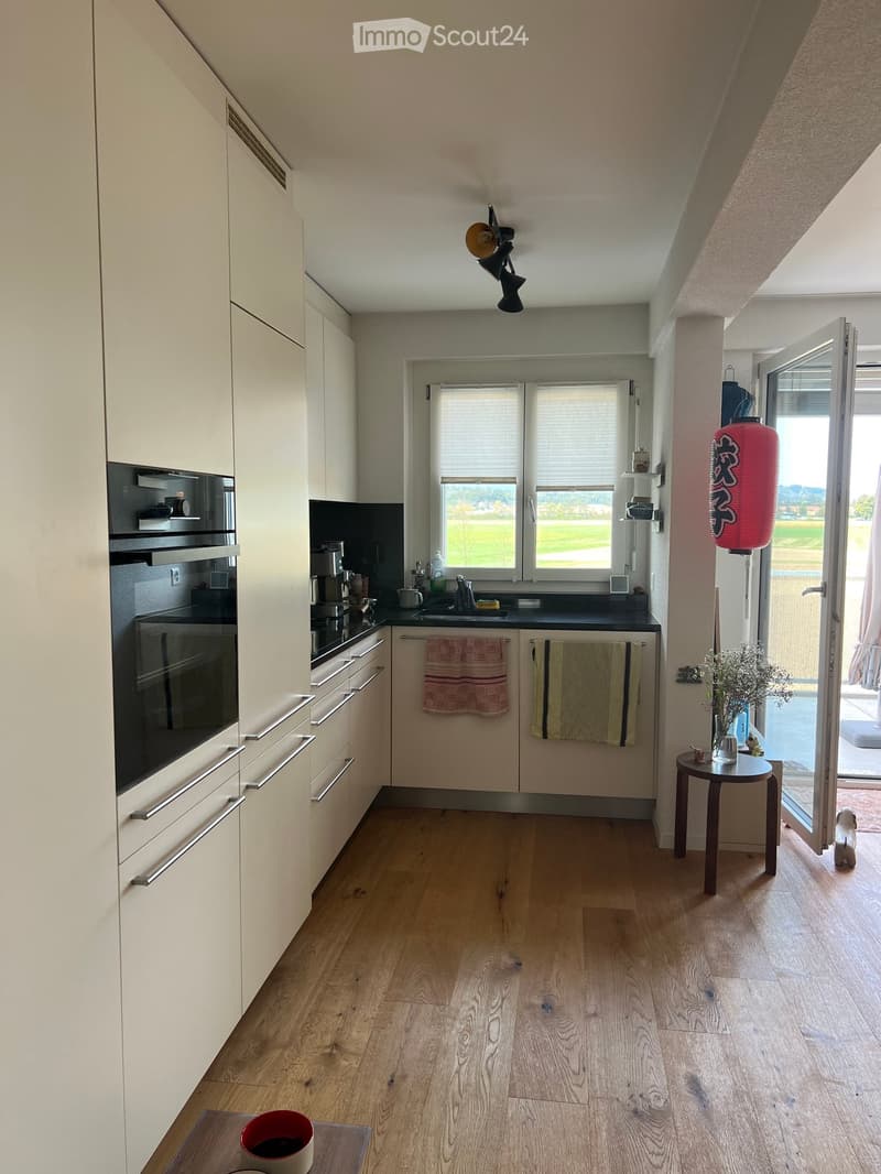 Modern kitchen with light-colored cabinets, wooden flooring, and stainless steel appliances. Features a coffee maker, sink, and a small table with a cup and a table lamp.