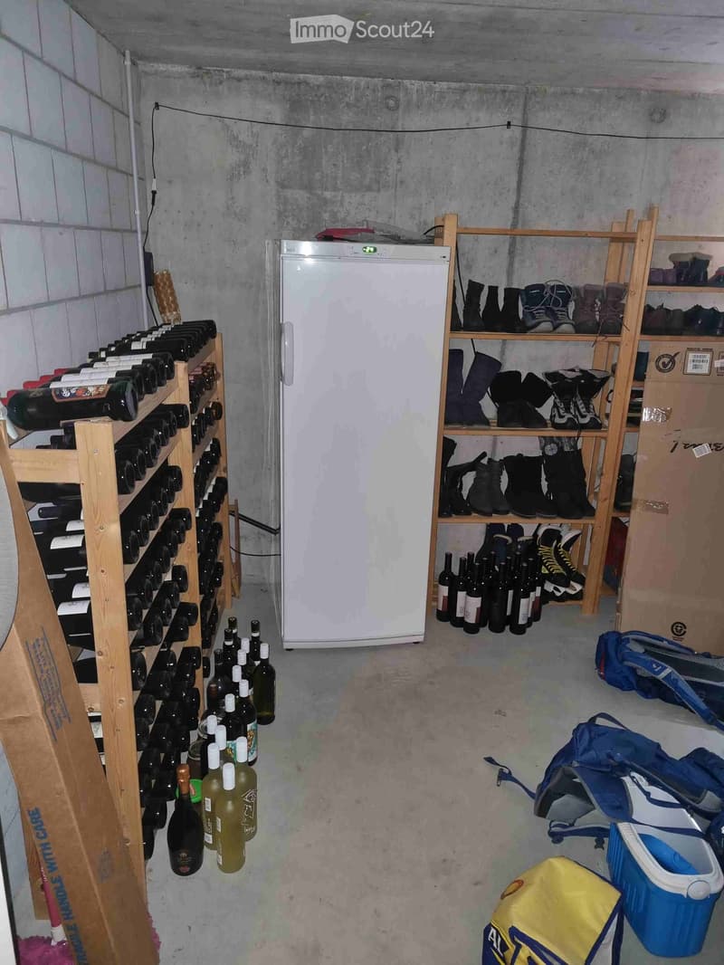 A small, concrete-walled storage room with a refrigerator, wooden wine racks, shelves holding shoes and bottles, and various bags and boxes on the floor.