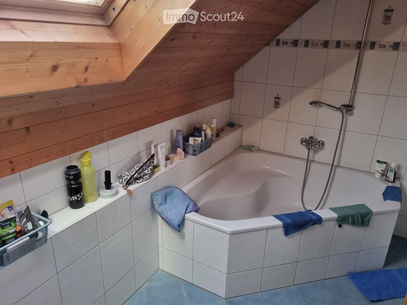 Bathroom with a white bathtub, tiled walls, wooden slanted ceiling, and various toiletries on the edge of the tub.