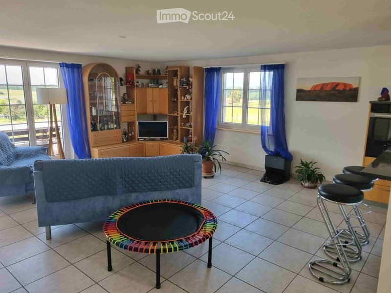 Bright living room with large windows, blue curtains, a blue sofa, a colorful round coffee table, bar stools, a wooden cabinet, and a television. The room features tiled flooring and potted plants.