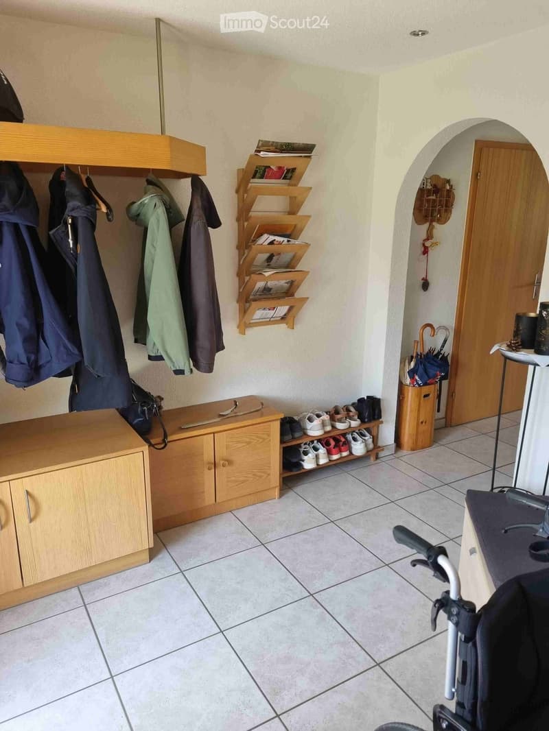 A well-organized hallway with wooden coat rack, shoe rack, umbrella stand, and storage shelves. The space features tiled flooring and a small wooden bench with a drawer.