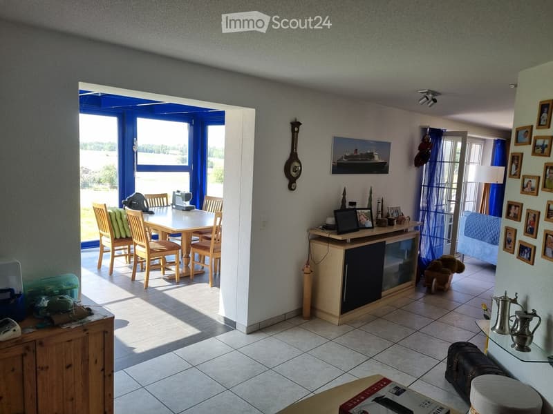 Bright living area with a dining table and chairs, a small cabinet with a laptop, picture frames on the wall, and a view through large windows.