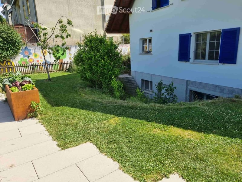 Small garden area with a paved walkway, grassy lawn, and a raised planter box with vegetables. The garden is bordered by a wooden fence and a neighboring house with blue shutters.