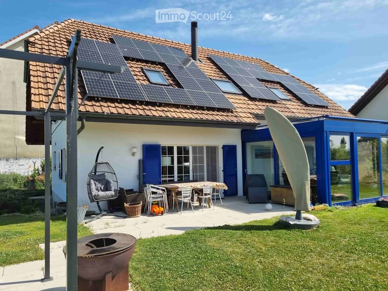 Garden with a patio area featuring a table, chairs, a wicker hanging chair, a grill, and a fire pit. The house has solar panels on the roof and blue shutters.
