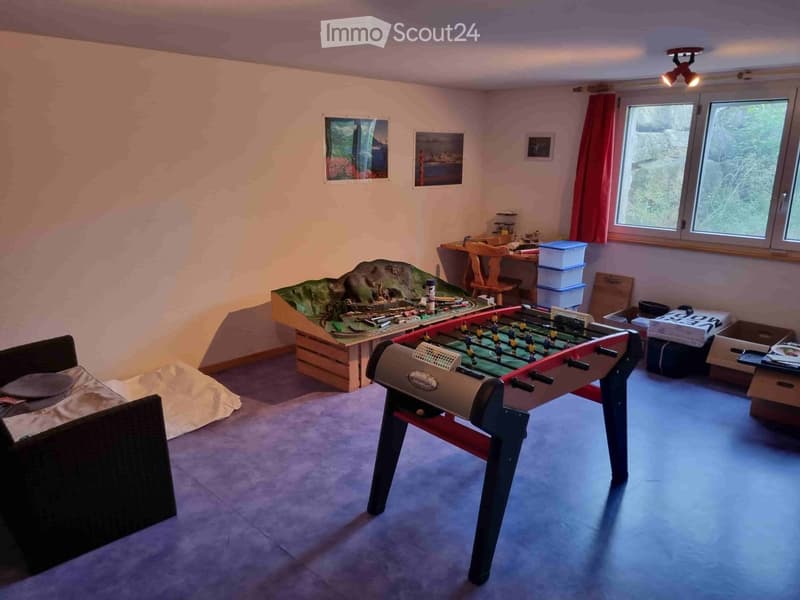 A cozy room with a sofa, futon, train set, foosball table, and storage boxes. The room has large windows with red curtains and is well-lit with ceiling spotlights.