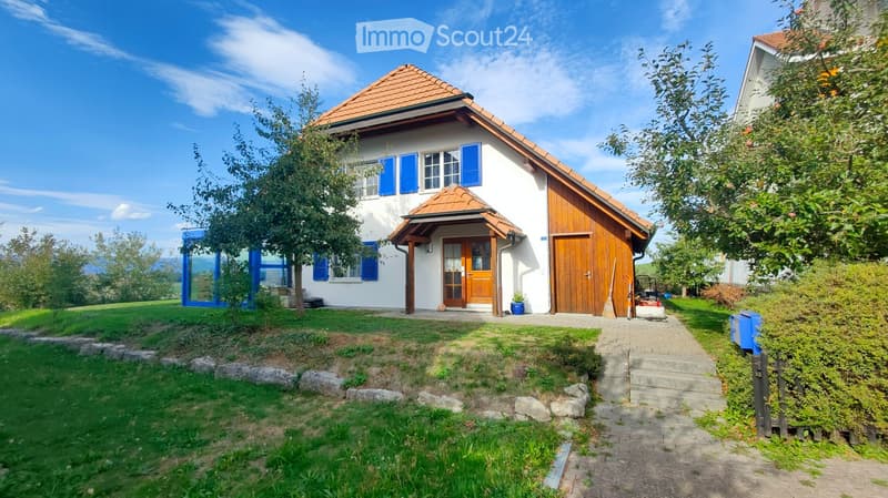 Two-story house with white walls, blue shutters, and a red-tiled roof. Features a wooden garage, a small porch with a wooden door, and a landscaped front yard with a stone pathway.