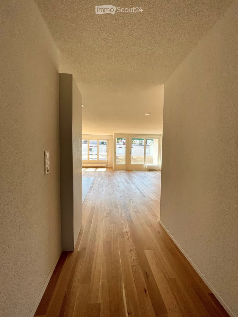Empty hallway with wooden flooring, light-colored walls, and recessed ceiling lighting. The hallway leads to a room with large windows and a sliding glass door.