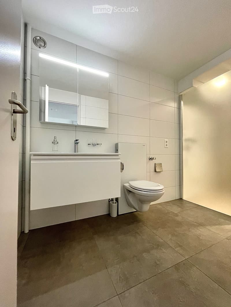Modern bathroom with white tiles, a sink with a modern faucet, a large mirror with built-in lighting, and a toilet. The floor is tiled in a neutral tone.