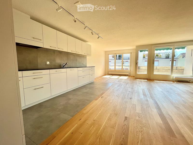 Empty kitchen with white cabinets, wooden flooring, and large windows allowing natural light. No furniture or appliances are present.