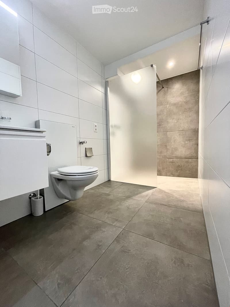 Modern bathroom with a toilet, sink, and cabinet, featuring light grey tiles and a glass shower enclosure.