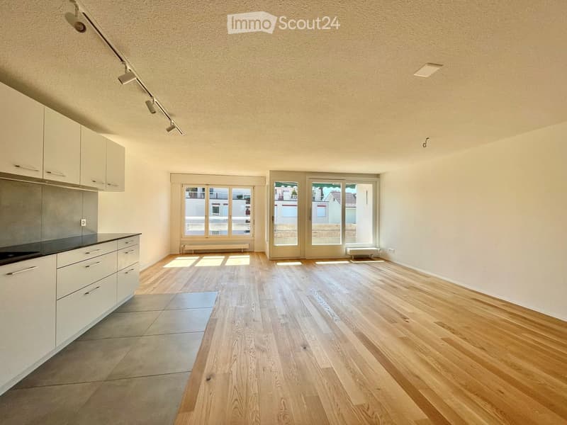 Empty room with light wooden flooring, white walls, and a small kitchen area with cabinets and a countertop. Large windows and sliding glass doors lead to a balcony.