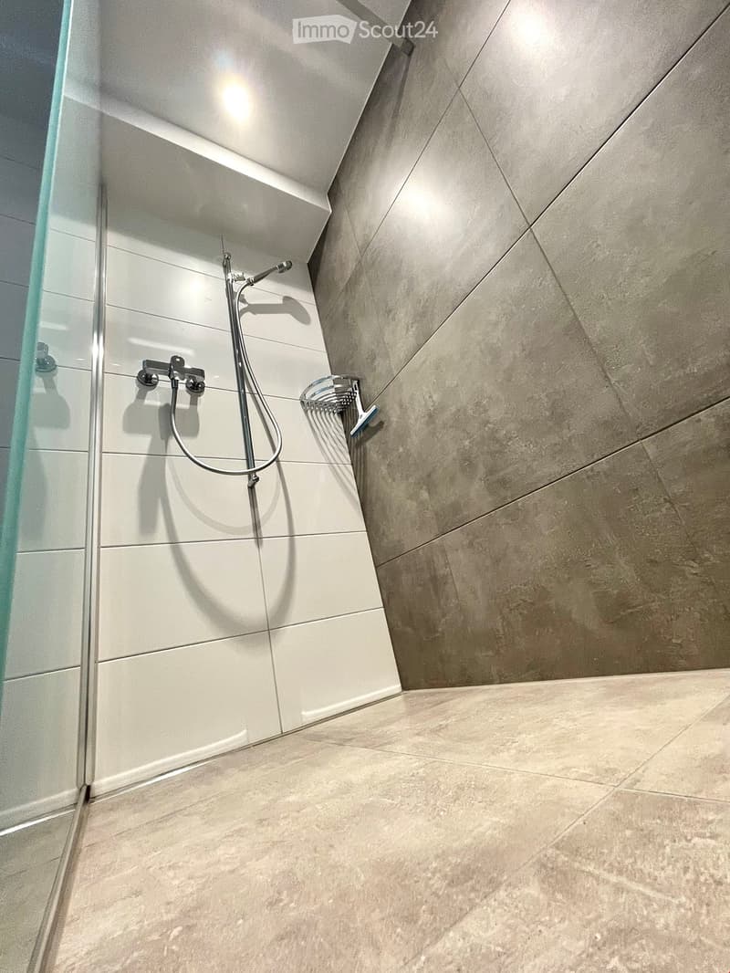 Modern bathroom with a glass-enclosed shower area, featuring a sleek showerhead and faucet, light grey tiles on walls and floor, and recessed lighting.