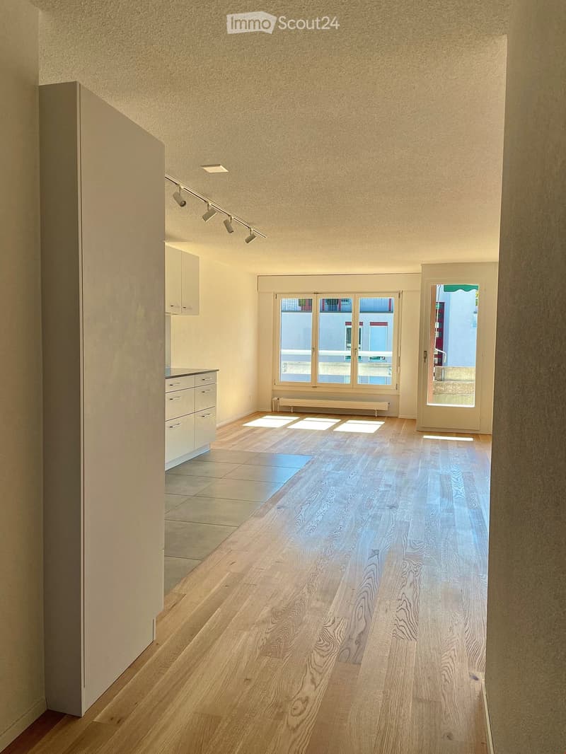 Empty hallway with wooden flooring, white walls, and recessed lighting. Features a small cabinet and a window with a view of the outside.
