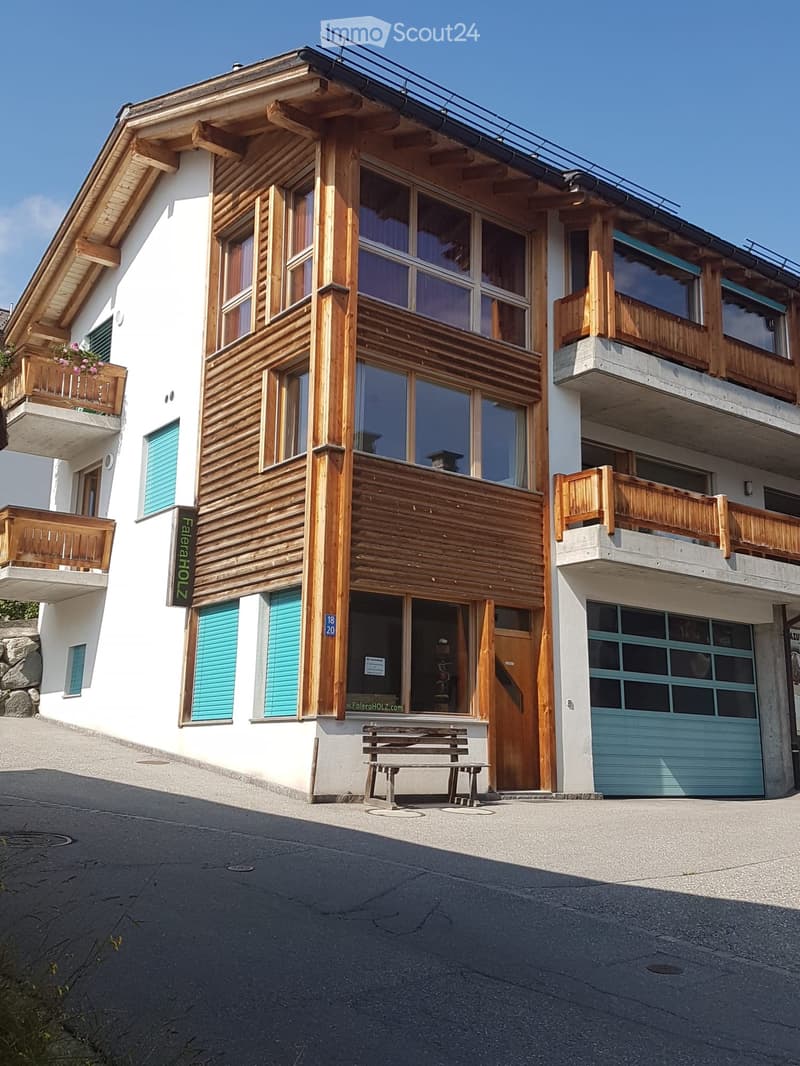 Two-story wooden building with balconies, large windows, and a garage. The building has a white facade with wooden accents and a slanted roof.