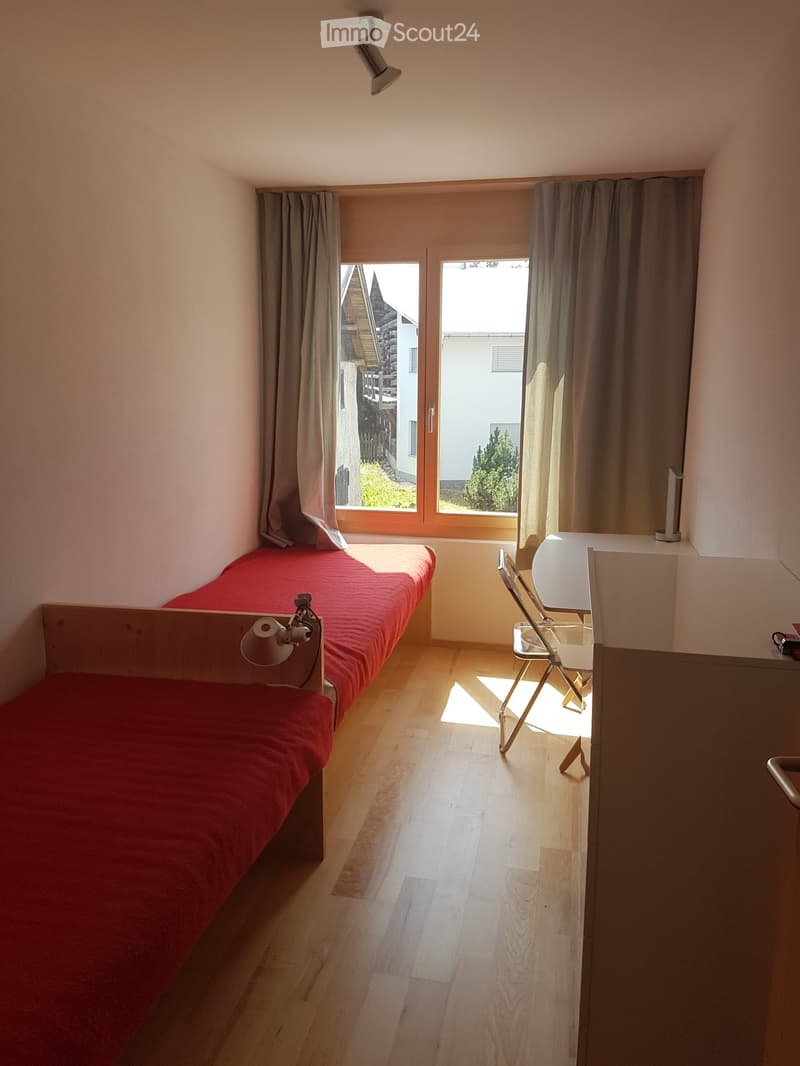 Cozy bedroom with a single bed, a small table and chair, and large windows with curtains.