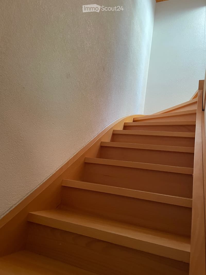 Wooden staircase with light-colored steps and railings, set against a textured beige wall and white upper wall.
