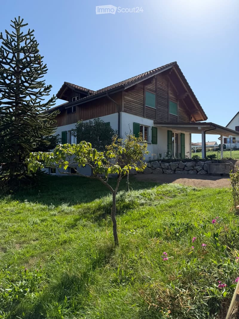 Two-story house with wooden exterior, green shutters, and a stone wall in front. The house is surrounded by a well-maintained lawn, small trees, and shrubs.