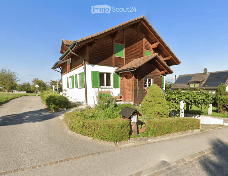 Two-story house with wooden and white exterior, green shutters, and a well-maintained garden with bushes and trees.