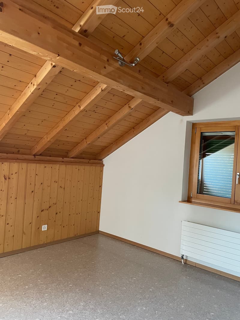 Empty attic space with wooden slanted ceilings, exposed beams, concrete floor, white walls, and a single window with blinds.