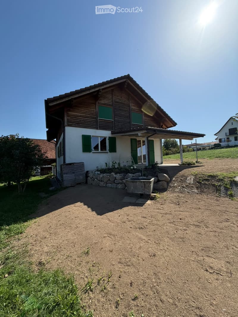 Two-story house with white walls, green shutters, wooden roof, and stone foundation. The house has a covered entrance and is surrounded by a dirt driveway and grassy areas.