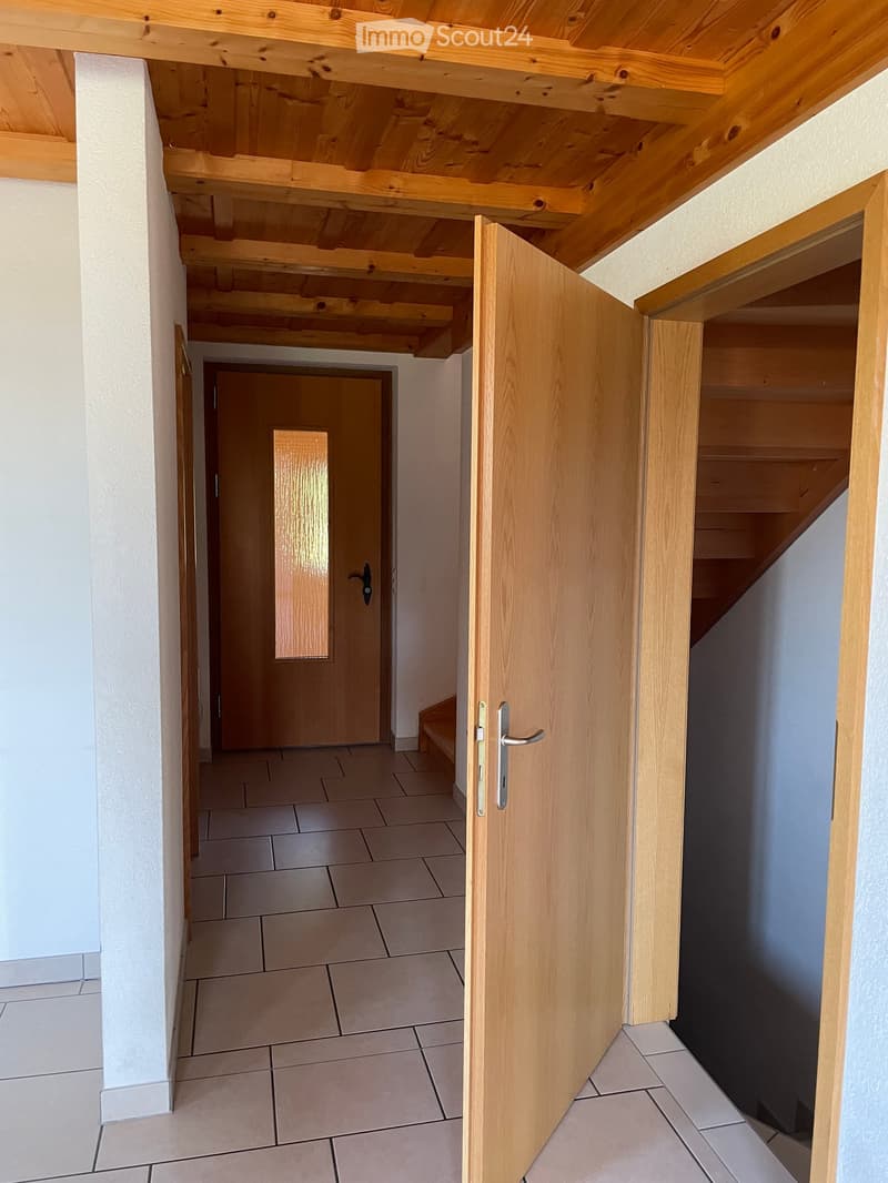A narrow hallway with tiled flooring, white walls, and wooden doors. The hallway leads to a staircase with wooden railings and a visible wooden ceiling structure.
