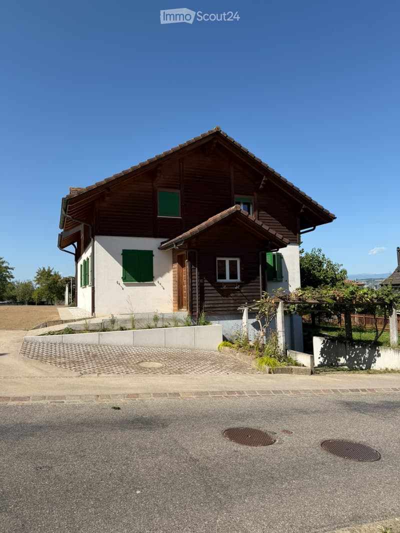 Two-story house with wooden facade, green shutters, and a tiled roof. The house features a raised ground floor and is surrounded by a low concrete wall and some greenery.