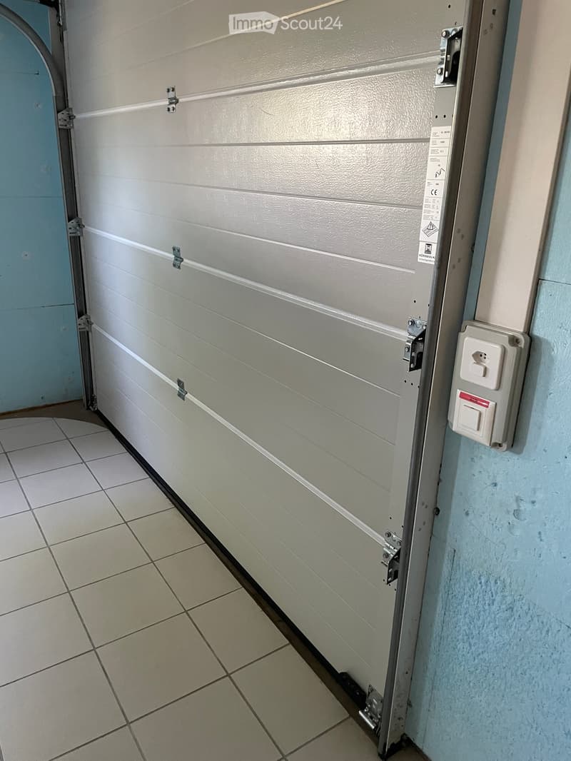 A partially open garage door with a tiled floor and control panel visible on the wall.