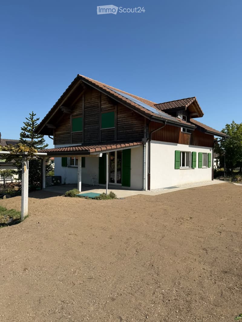 Two-story house with wooden facade, green shutters, solar panels on the roof, and a covered entrance area.