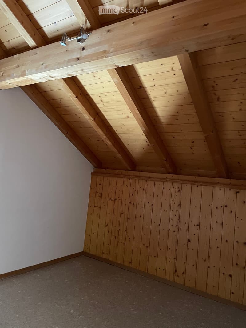 Empty attic space with exposed wooden beams and slanted wooden ceiling, grey carpeted floor, and white painted wall.