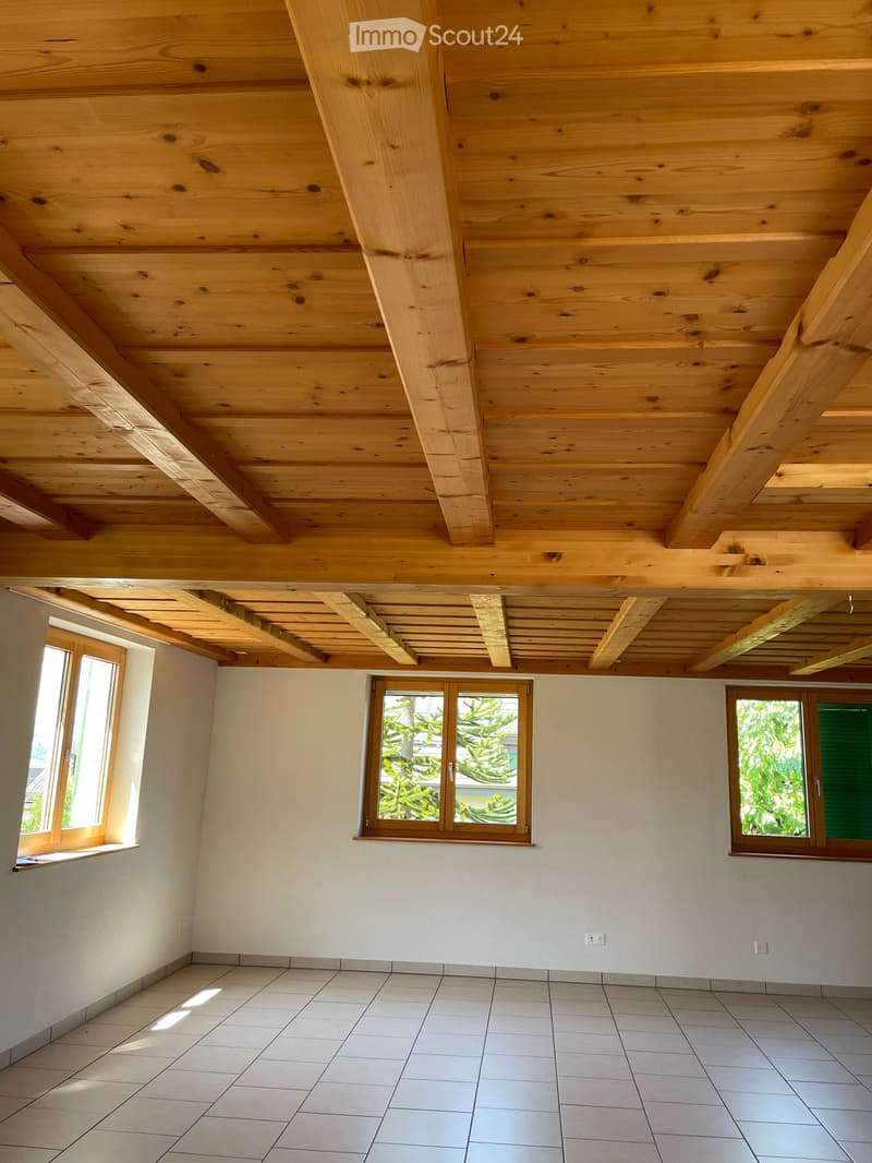 An empty room with exposed wooden beams on the ceiling, tiled floor, and three windows with wooden frames. The walls are painted white, and the room has a clean, minimalist design.