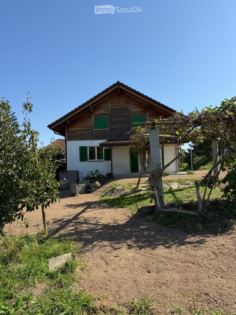 Two-story wooden house with green shutters, surrounded by trees and a gravel pathway.
