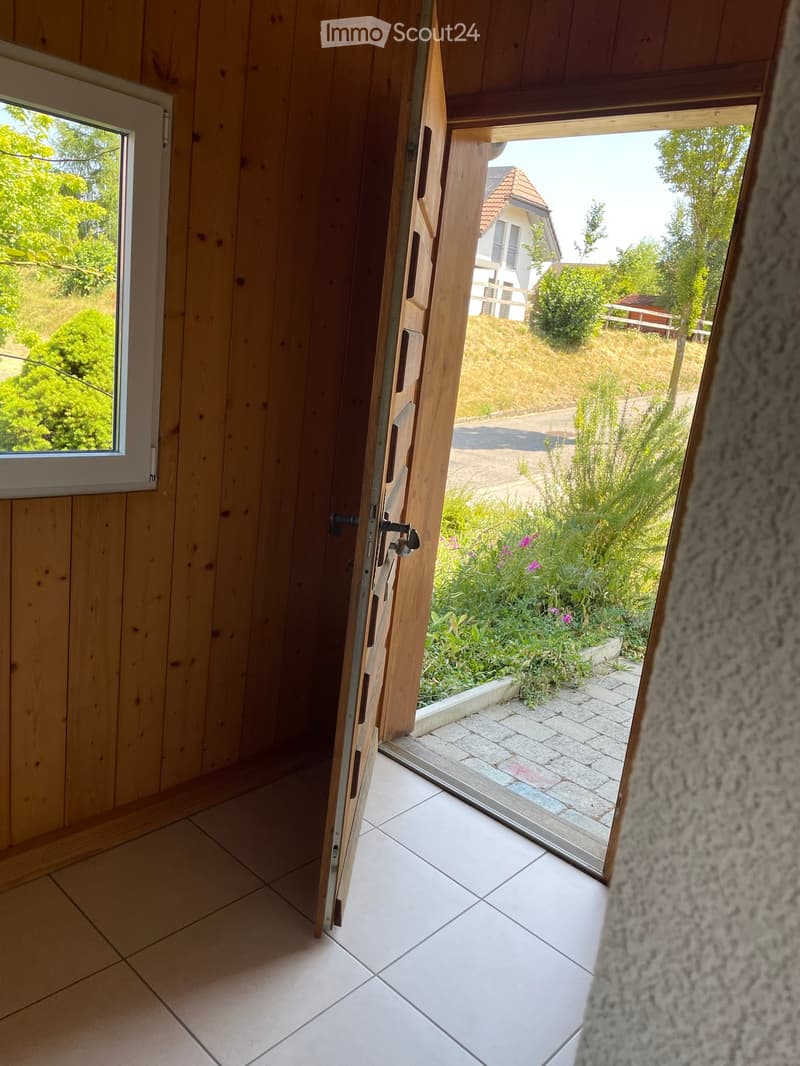 Open doorway leading to a garden area with plants and a paved pathway, visible from inside a wooden-walled corridor.