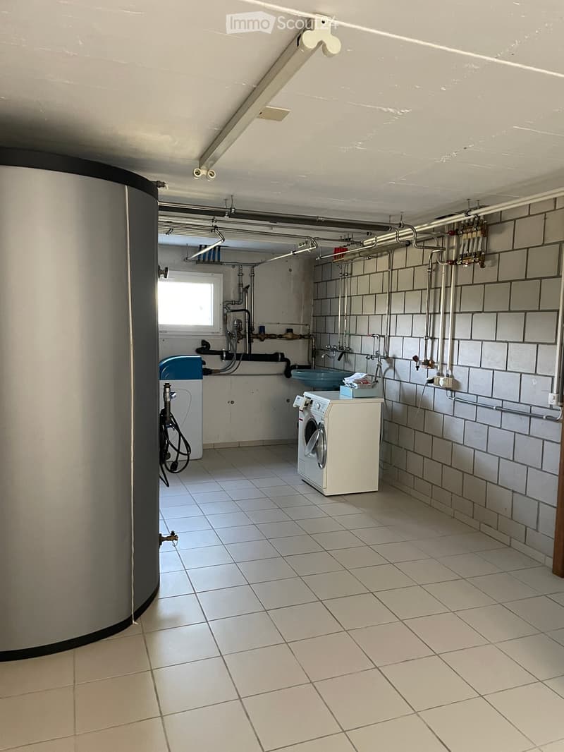 Utility room with tiled floor, white washing machine, sink, and water tank. Exposed pipes and valves visible on walls.