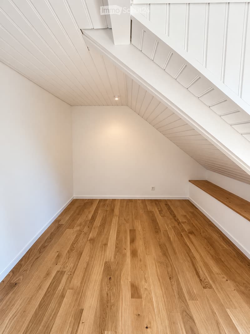 Empty room with wooden flooring and white walls, featuring a slanted ceiling and built-in wooden bench along one wall.
