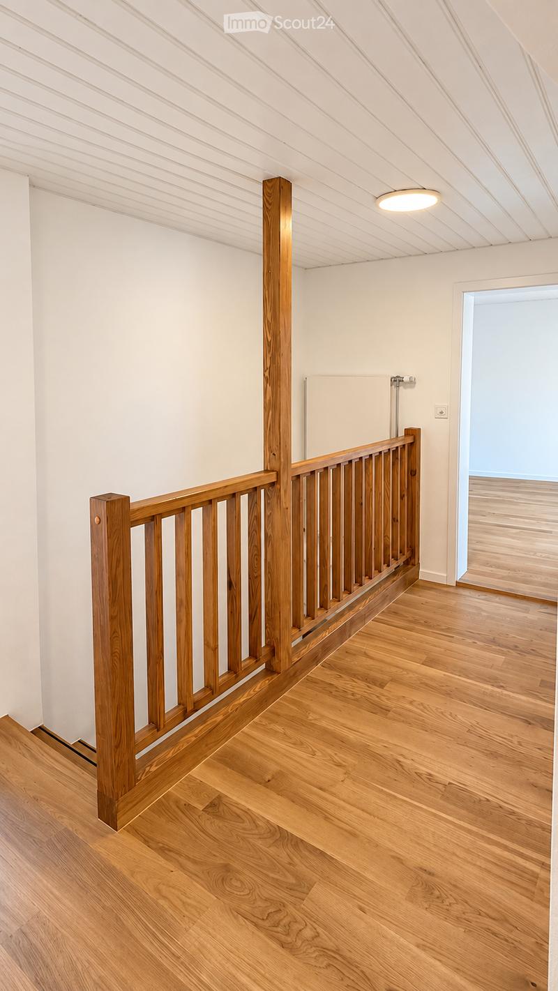 Spacious hallway with wooden flooring, white walls, and a wooden railing leading to another room. A recessed ceiling light illuminates the area.