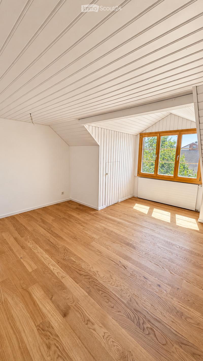 Empty room with wooden flooring, white walls, and a large window with wooden frames. The ceiling has exposed wooden beams and panels.
