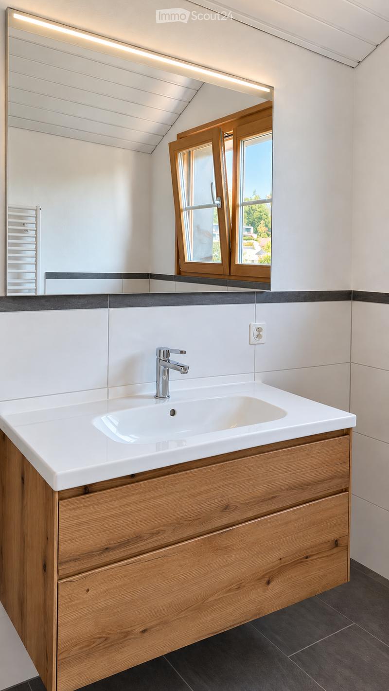 Modern bathroom with a white sink, wooden cabinet, chrome faucet, and large mirror above the sink. The room features dark tiled flooring and a window with wooden frames.